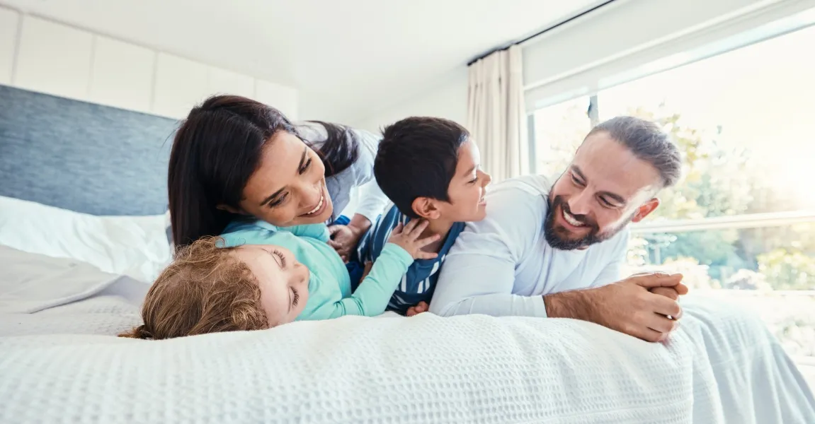 A happy family of four smiling and relaxing together on a clean, white bed, enjoying peace of mind in a pest-free home.