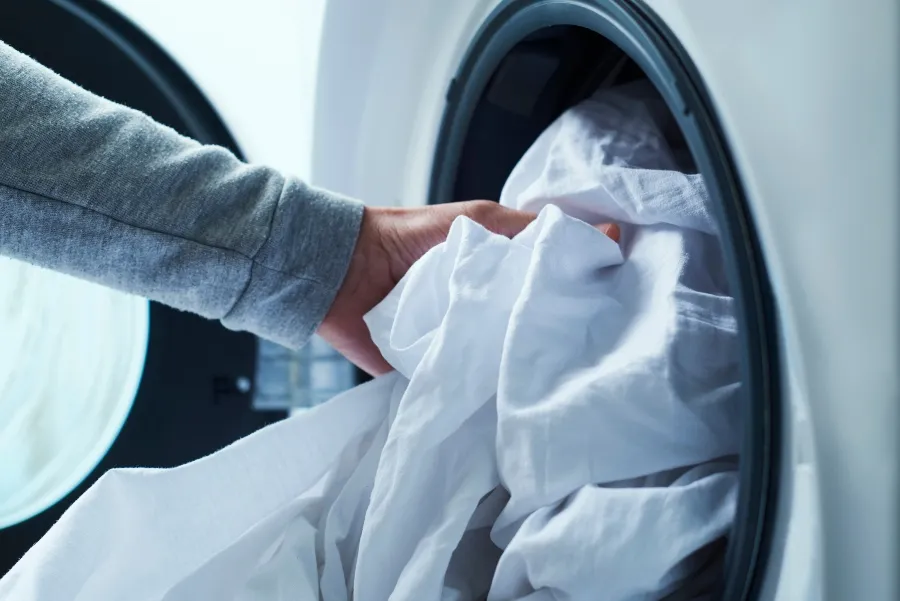 A person loading white bed sheets into a washing machine on a high heat cycle as a step in DIY bed bug treatment.