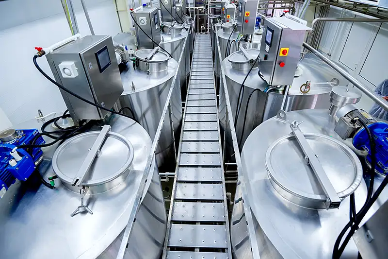 A modern food processing facility with a row of large stainless steel tanks connected by a central elevated walkway. The tanks are equipped with control panels, pipes, and sealed lids, showcasing an industrial setup for handling or manufacturing food products in a clean and controlled environment.