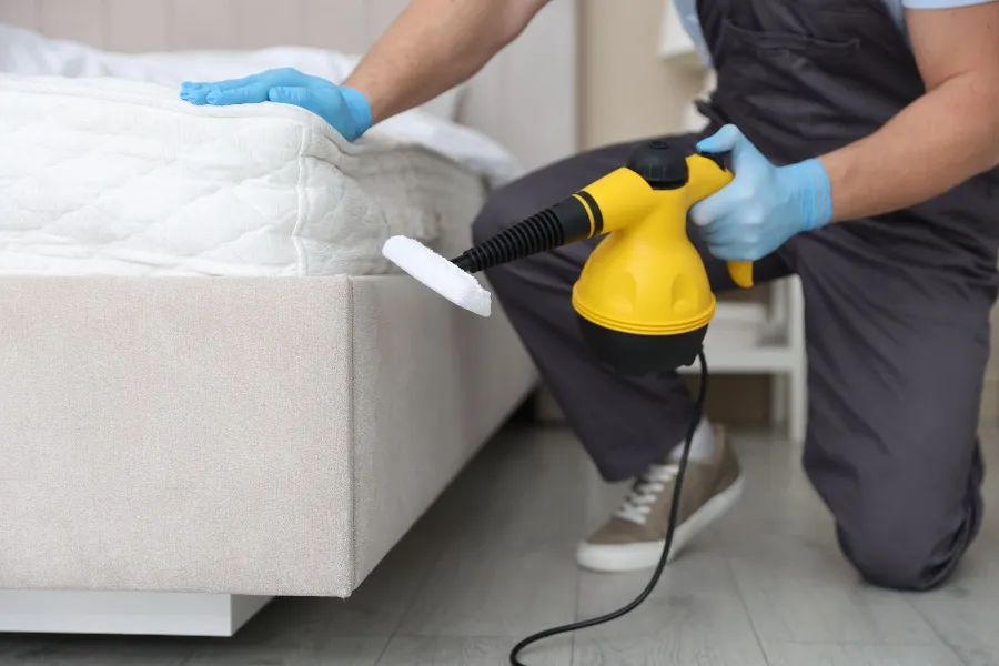 A pest control worker using a yellow handheld steam cleaner on a mattress to prevent and eliminate bed bugs.